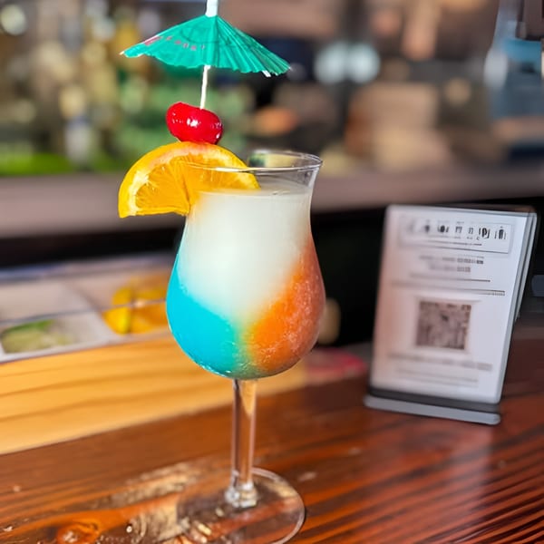 A colorful frozen cocktail with orange, blue, and white layers, garnished with an orange slice, cherry, and umbrella.