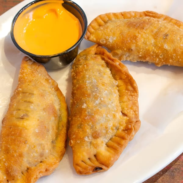 Three golden-brown empanadas and a small black cup of orange dipping sauce on a white plate.