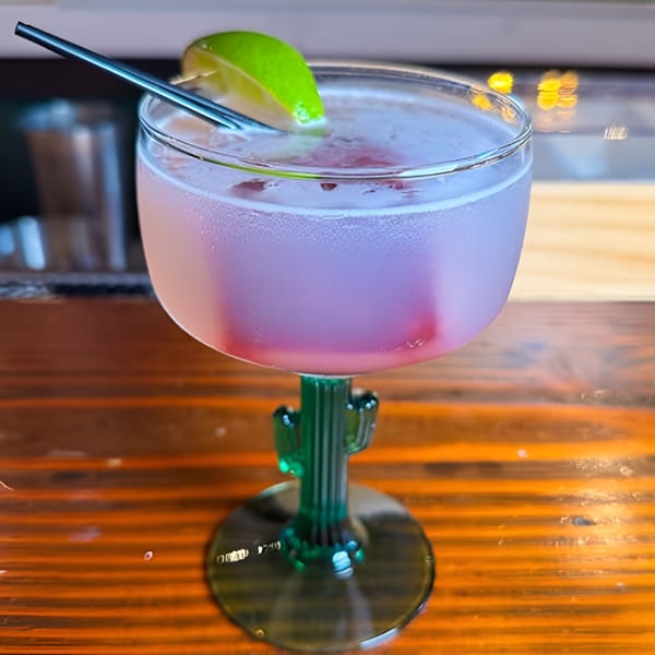 Pink margarita in a cactus-stemmed glass with a lime wedge and straw on a wooden bar.